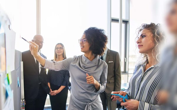 A woman speaks to a group dressed in business attire in front of a whiteboard in an office.