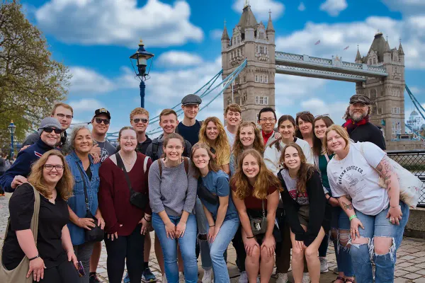 A group of students pose in front of London Bridge with a blue, cloudy sky.