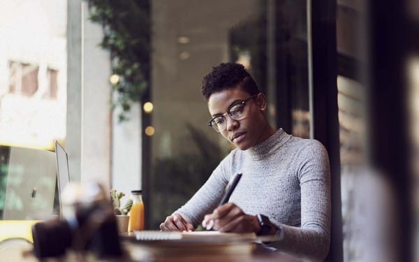 A woman sits at a desk in front of an open laptop as she writes in a notebook.